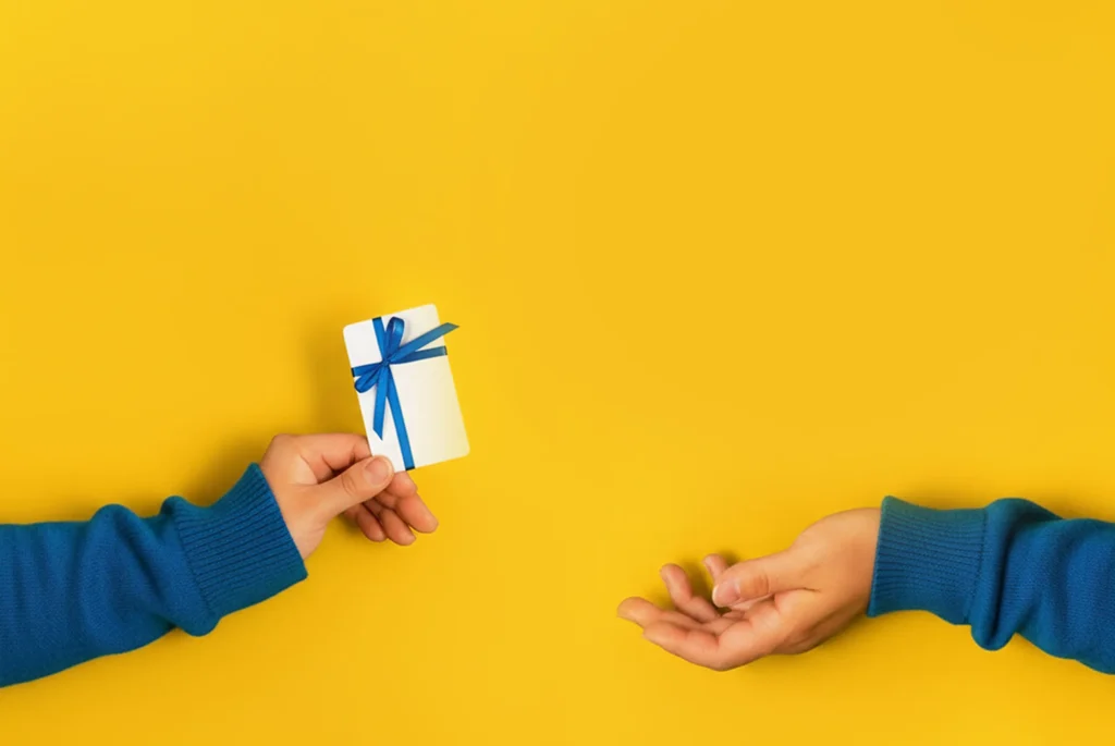 Mano che consegna una carta regalo con fiocco blu su sfondo giallo, simbolo delle Gift Card Digitali disponibili su APay E-Wallet.